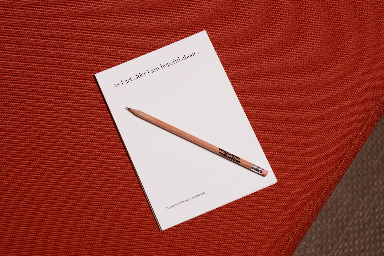 A small white booklet lies on a red textured surface, with a pencil resting diagonally across the cover. The booklet is titled “As I get older I am hopeful about...” and invites the viewer to draw or write a response.