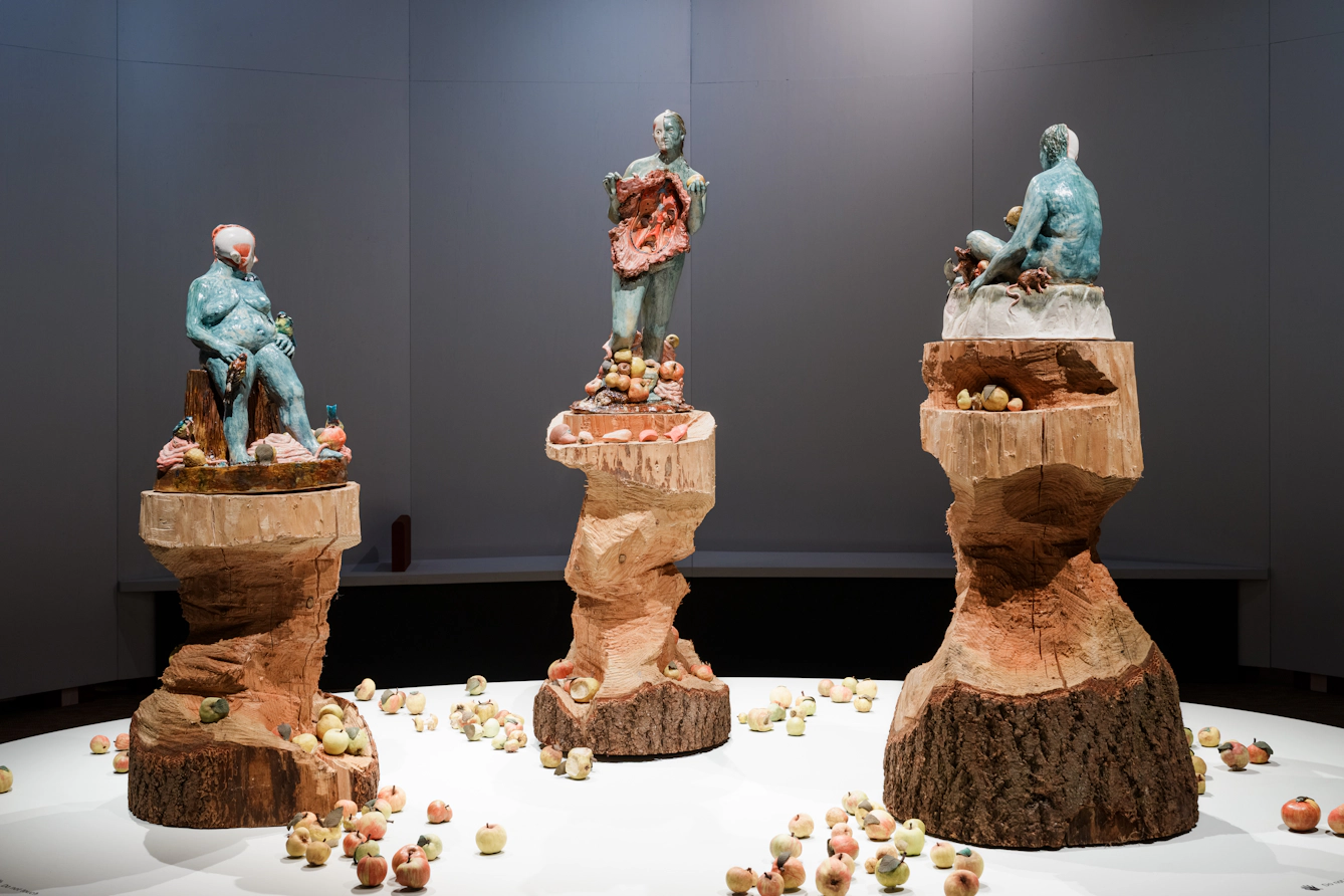 Sculptures of three women on large wooden pillars, surrounded by apples. The central figure's body has been opened up and her insides are visible. 