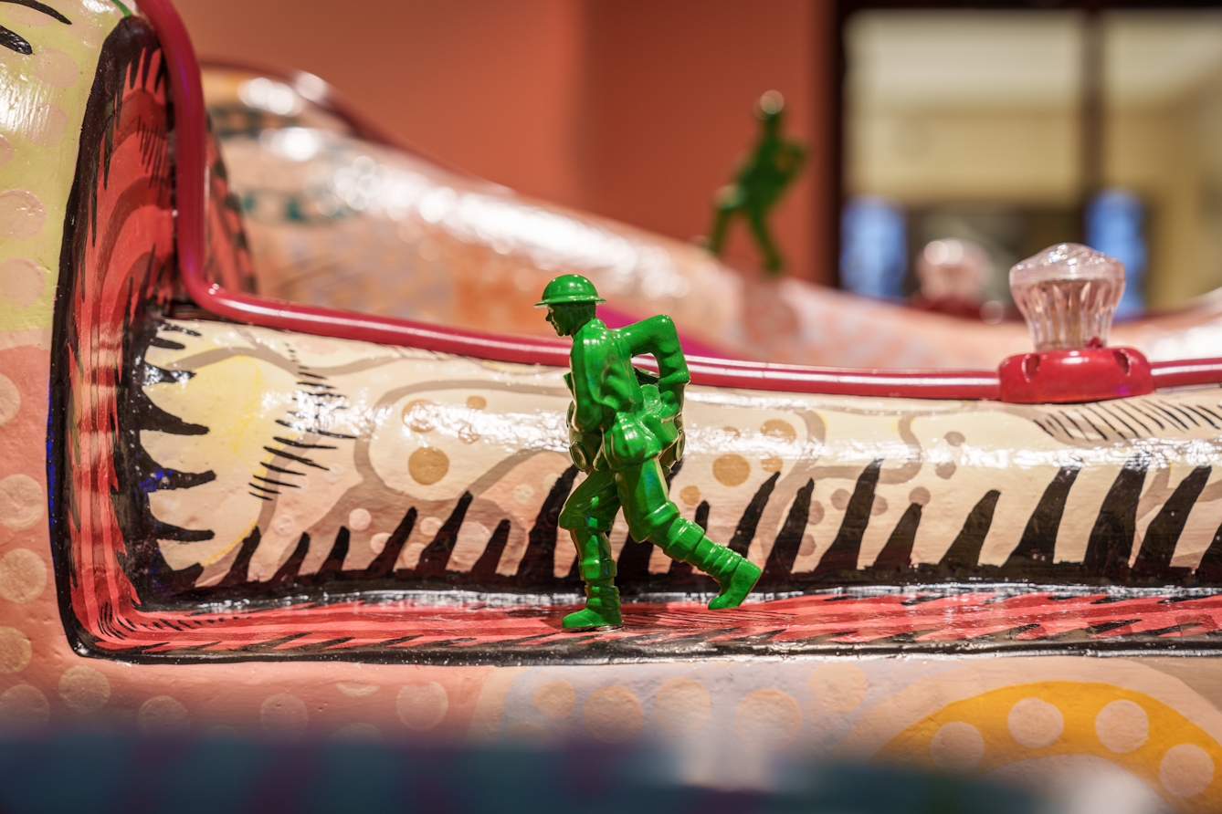 Detail from a larger sculpture showing a cut away section of the sculpted body's leg, and a small green toy soldier marching up the cut away.