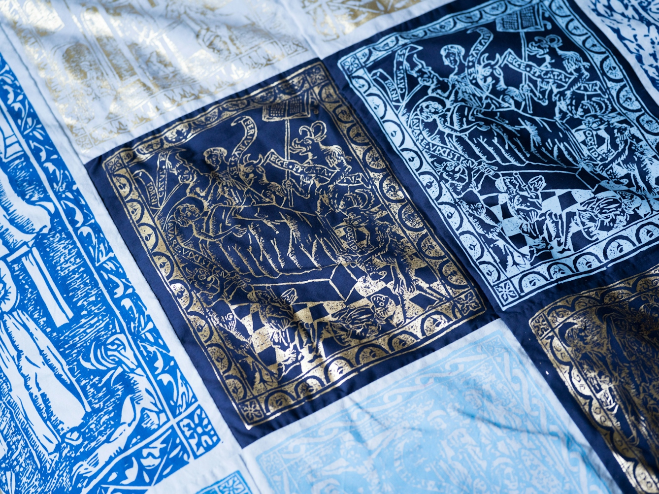 Photograph of a close up of a duvet cover which is made of a patchwork of blue, silver and gold screen-printed designs. 