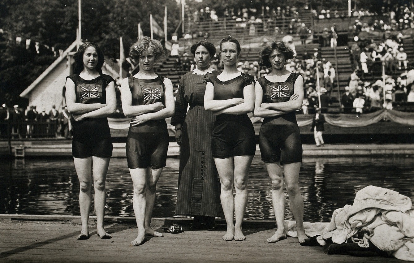 Black and white photo of five women standing in a line in front of a pool. Four of them wear old-fashioned swimming costumes. The other one wears a long dress.