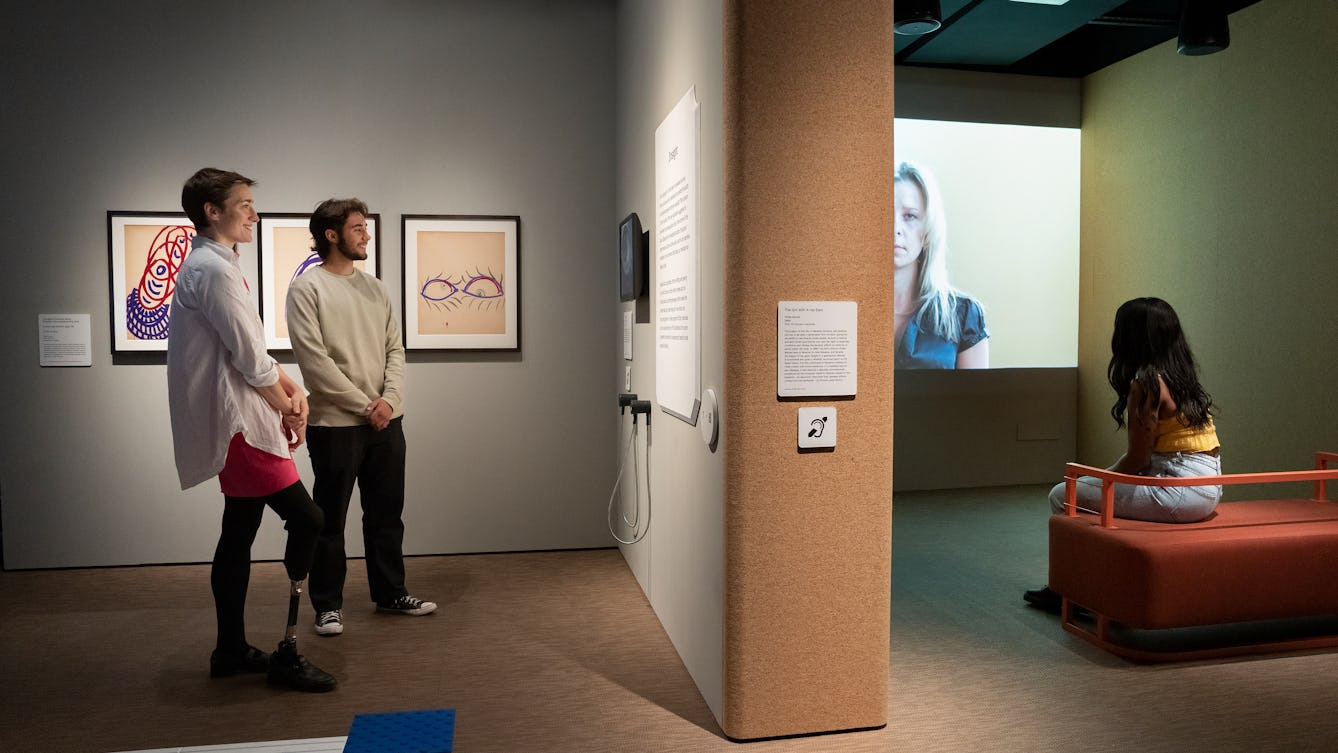 Photograph of people exploring an exhibition. Two people are stood side by side looking at information on the wall. One person is seated watching a film projected on the gallery wall. 