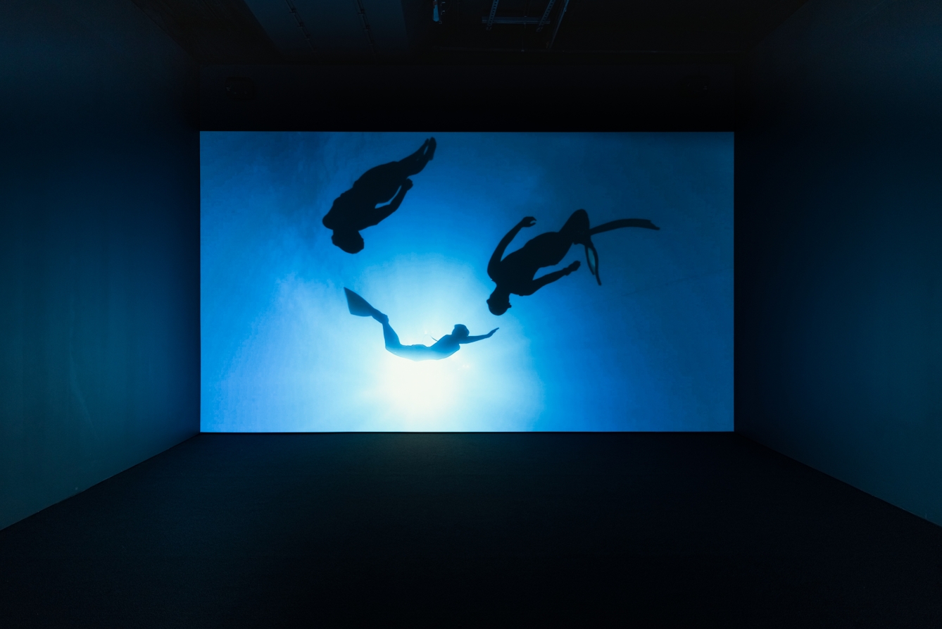 Photograph of a dark gallery with a large projection showing figures floating in water against one wall, which casts blue light into the room.