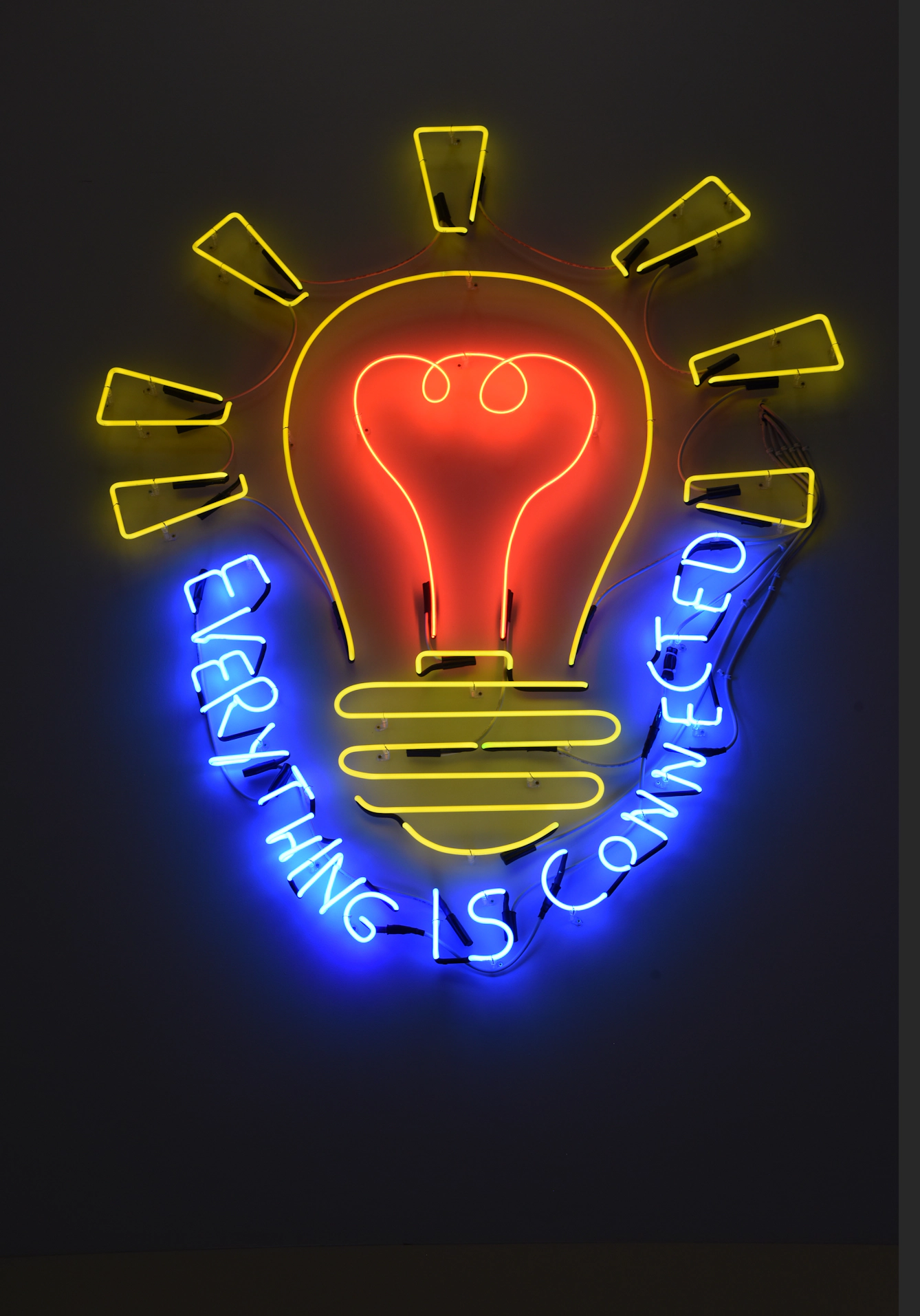 A neon sign shaped like a lightbulb glows against a dark wall. Blue tubing spells out “everything is connected” around the bulb.