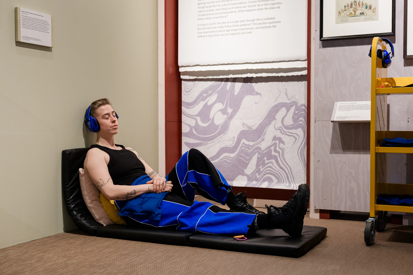 A visitor wearing ear defenders rests on a mat on the floor and leans against the wall.