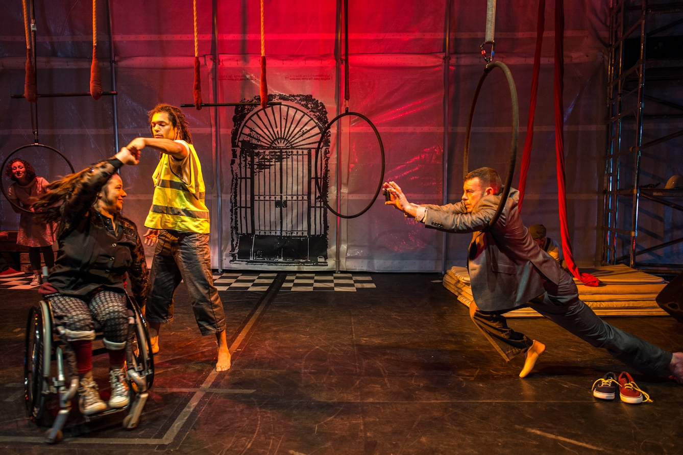 Photograph of a theatre stage with several performers. Stage left two performers, one standing and one seated in a wheelchair, perform a dance-like-manoeuvre where one twirls under the arm of the other. To the right of the frame a third performer is reaching towards the others through a large metal ring, as if his movements are restricted by the prop.  The stage is lit in a deep red hue.
