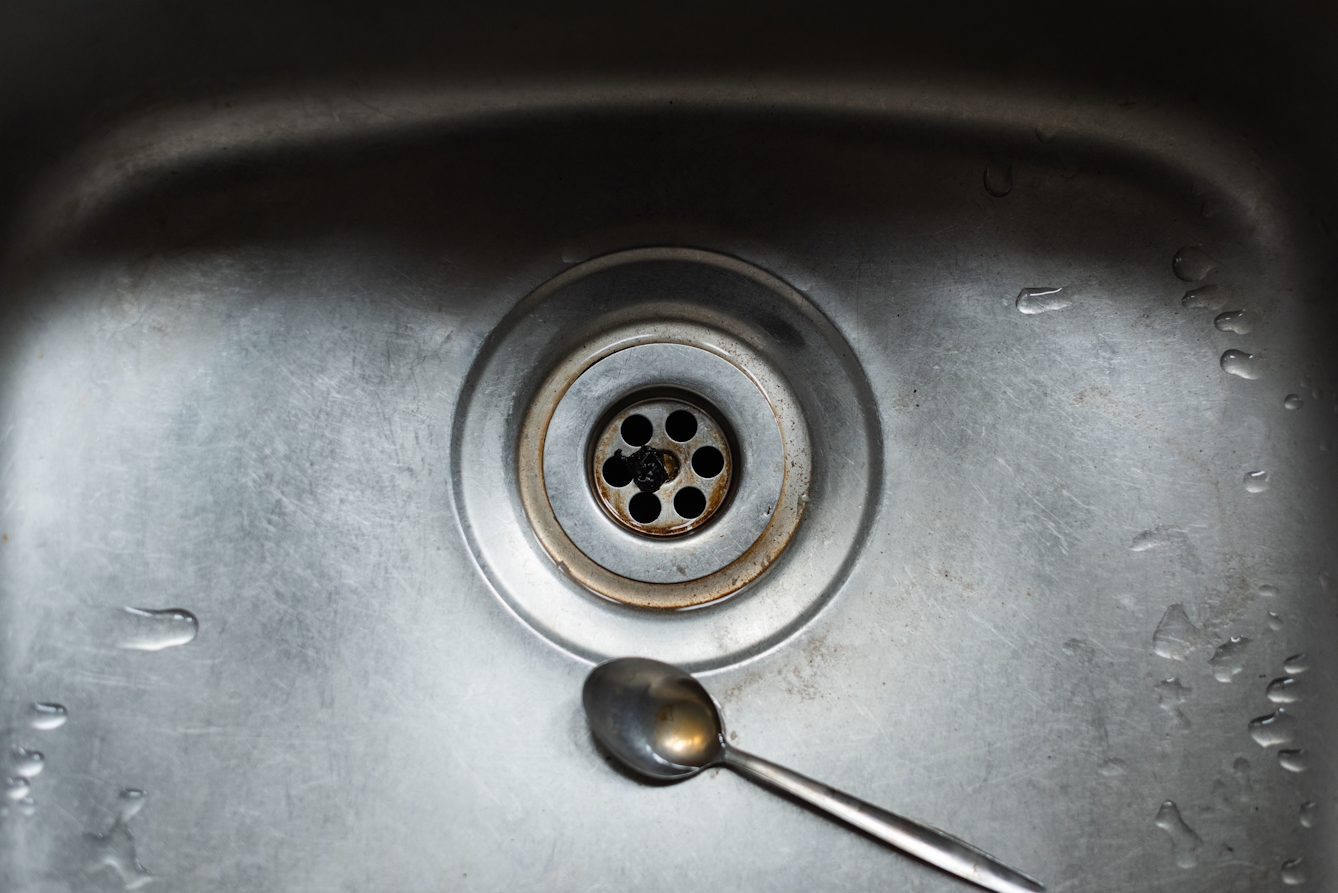 Photograph of a stainless steel kitchen sink, so close up that the sink fills the frame. In the centre is the plug hole with brown stains and a dark round squashed object. Next to the plughole is a teaspoon with a puddle of brown water in it. All over the surface of the sink are small patches of water.