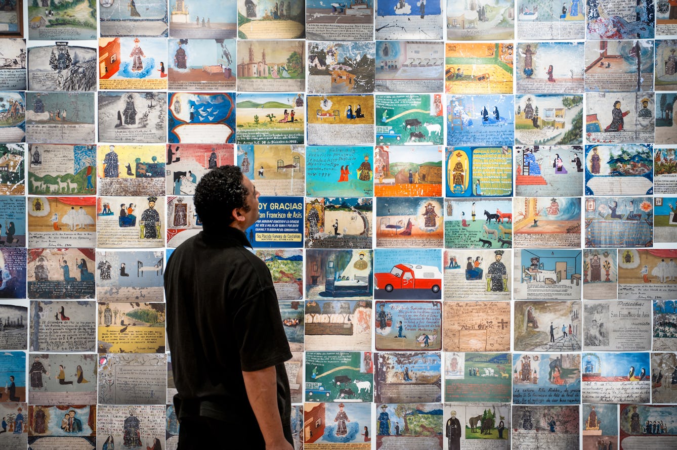 Photograph of an exhibition gallery wall covered in colourful Mexican votive plaques, depicting many and varied scenes. In front of the wall, stood in partial silhouette is a man, side on, look up at the votives.