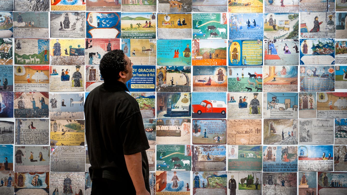 Photograph of an exhibition gallery wall covered in colourful Mexican votive plaques, depicting many and varied scenes. In front of the wall, stood in partial silhouette is a man, side on, look up at the votives.
