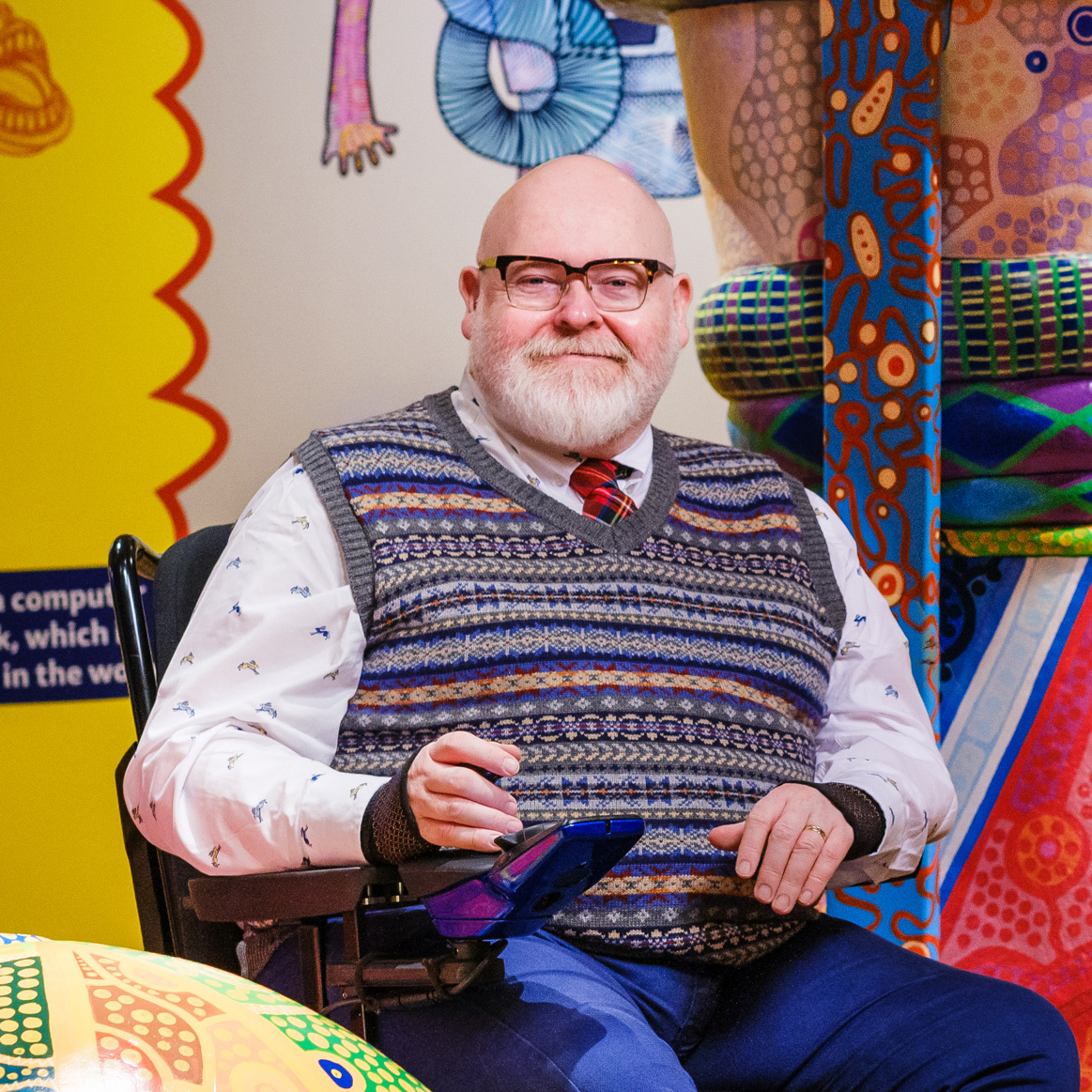 Photo of Jason Wilsher-Mills who is wearing a jumper vest over a shirt and tie with jeans