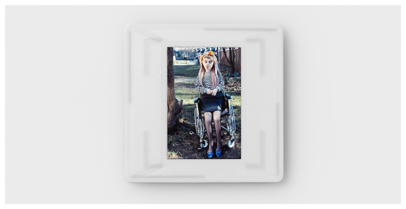 Photograph of a colour 35mm transparency mounted in a plastic side holder, resting on a white background. The transparency shows a young girl in a stripy black and white top, black skirt and blue shoes, sitting in a wheelchair. She has long hair, strands of which have been coloured blue, yellow and red. She also has a floral decoration clipped in her hair. She is pictured outdoors in a grassy space next to a tree trunk. In the distance behind her are a row of houses. 