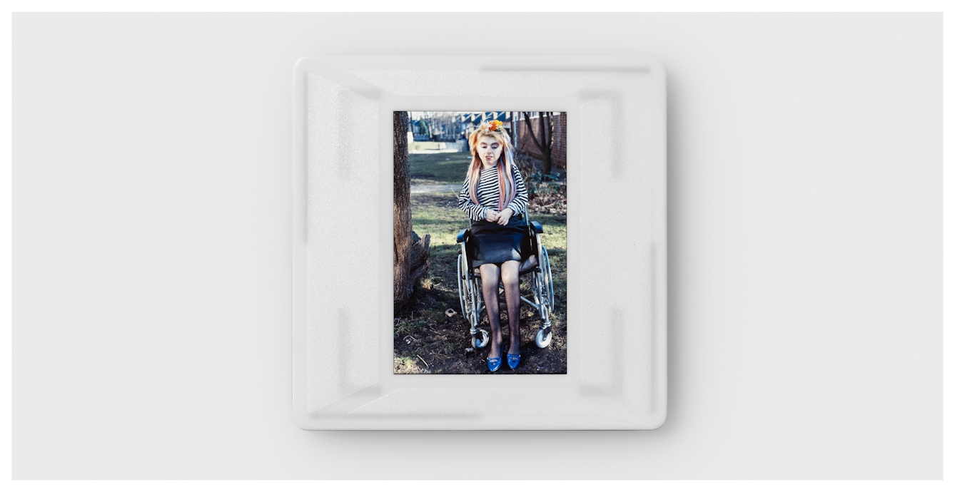 Photograph of a colour 35mm transparency mounted in a plastic side holder, resting on a white background. The transparency shows a young girl in a stripy black and white top, black skirt and blue shoes, sitting in a wheelchair. She has long hair, strands of which have been coloured blue, yellow and red. She also has a floral decoration clipped in her hair. She is pictured outdoors in a grassy space next to a tree trunk. In the distance behind her are a row of houses.