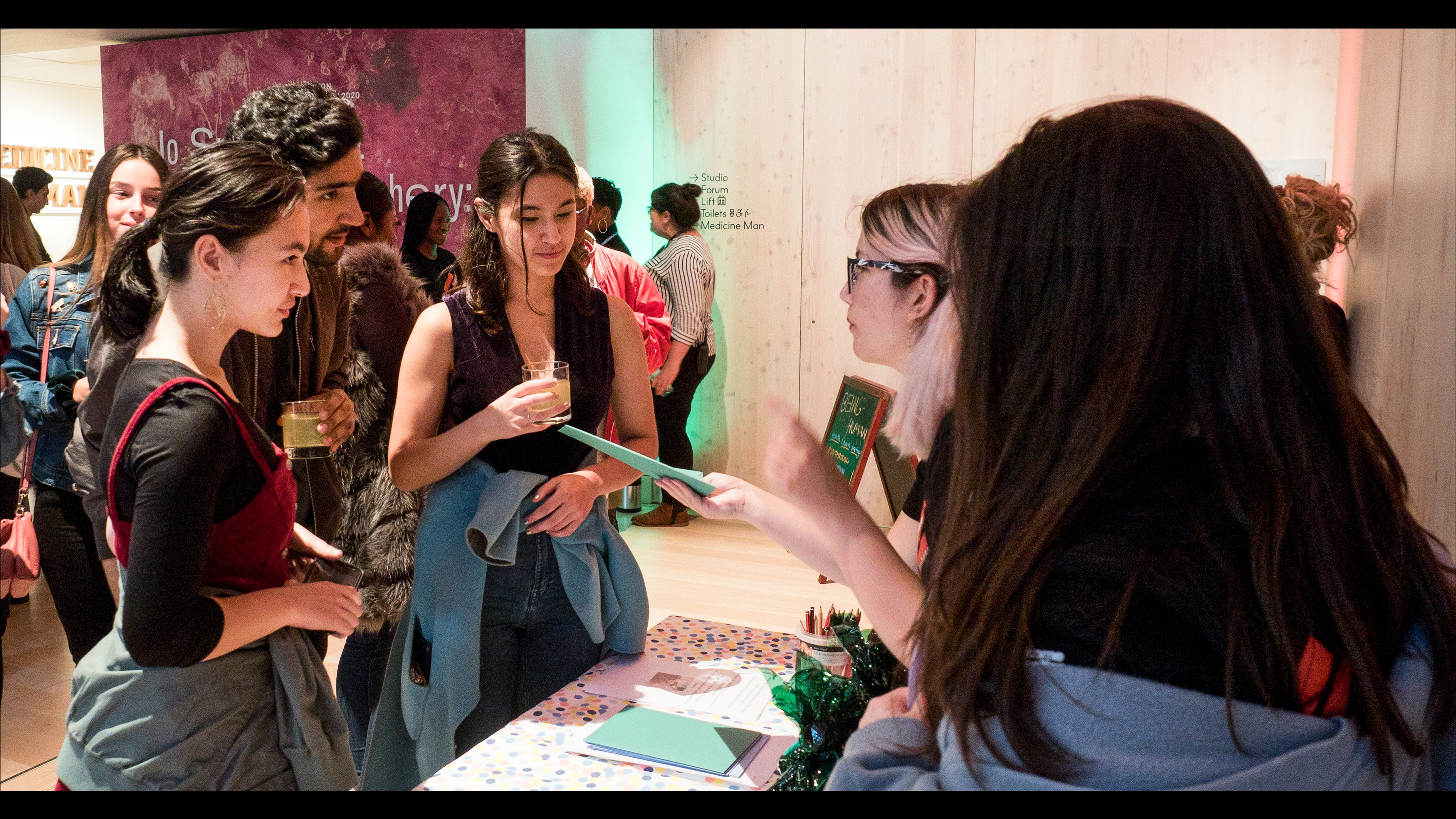 Young people standing outside the Being Human Exhibition at Wellcome Collection. That are holding mocktails and interacting with staff at a table who are handing  visitors paper exhibition guides.