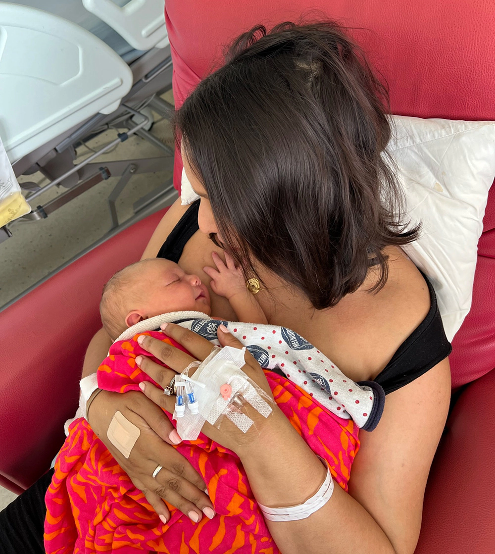 A mother sits in a chair looking down at the new-born baby in her arms. Her hands still are covered in tape and catheters from the birth.