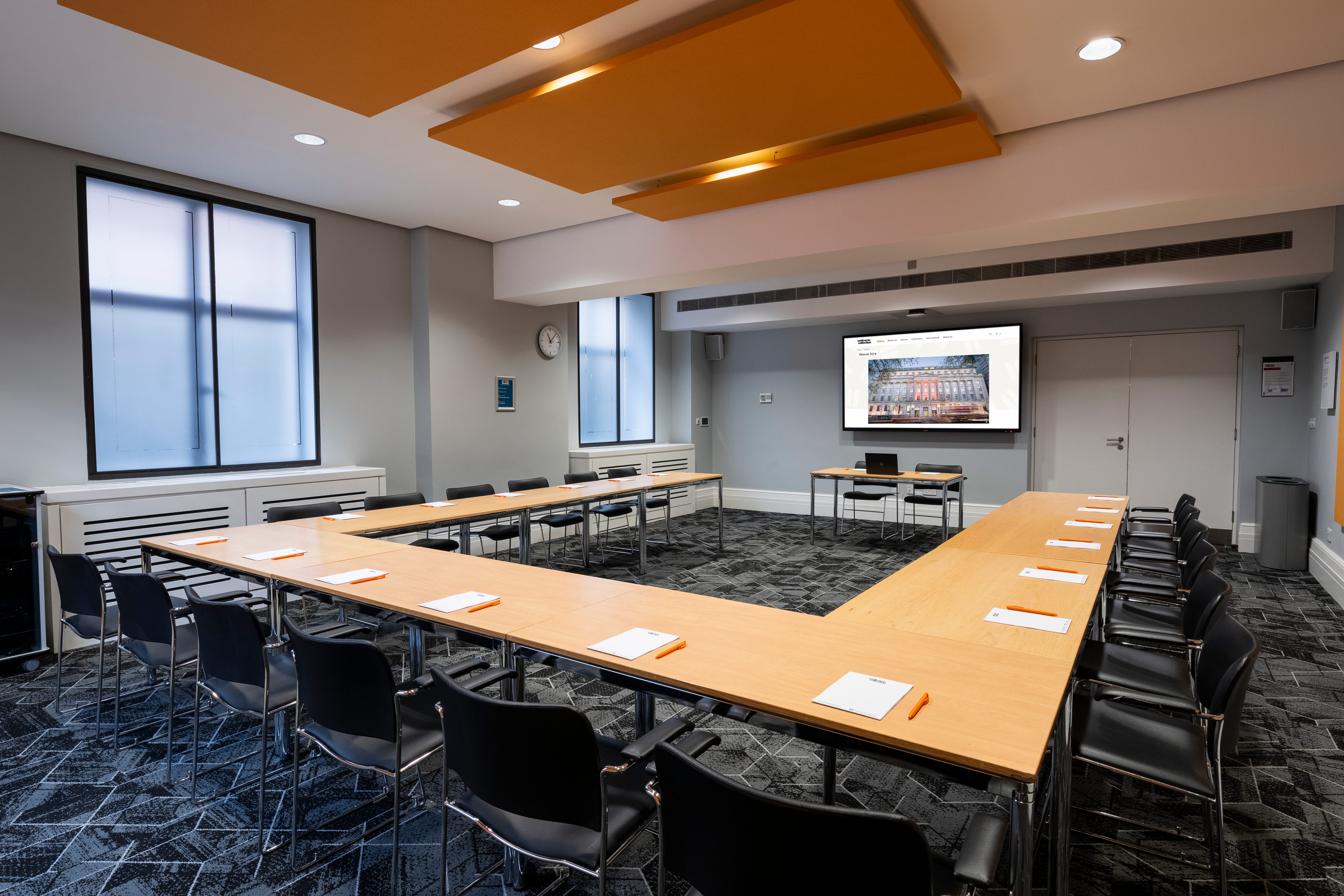 Meeting room with tables and chairs set with notepads and pens, a screen at the front, and windows letting in natural light.