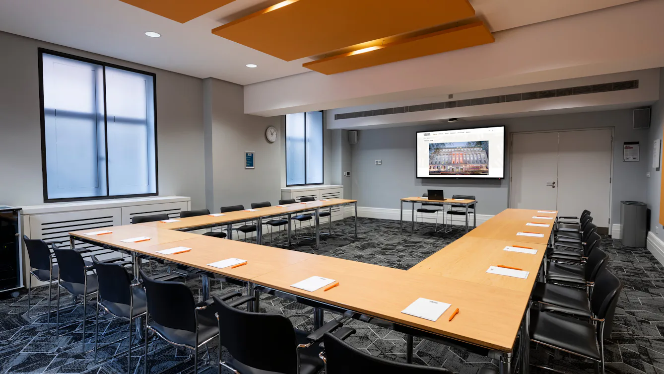 Meeting room with tables and chairs set with notepads and pens, a screen at the front, and windows letting in natural light.