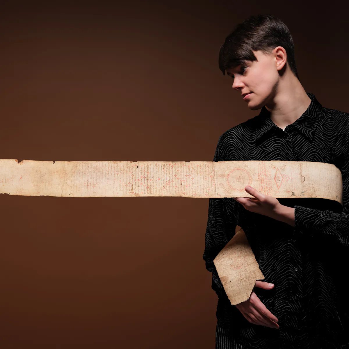Photographic portrait of Kierri Price with a 3 metre long facsimile of a medieval birthing scroll wrapped around their torso and then extending horizontally out of frame to the left. Kierri is in profile looking down at the hand written text and drawings which cover the scroll.