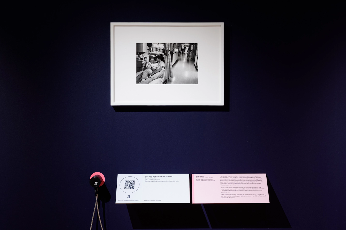 Photograph of a dark blue exhibition gallery wall showing a framed black and white photograph in a white frame. The photograph shows a hospital ward, with two people in casual clothes lying together on a narrow hospital bed surrounded by medical equipment, drinks and personal items, including photos and cards on the wall. Below the print is a caption panel and an audio guide ear cup.