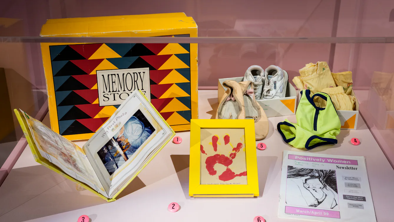 Photograph of an exhibition display case containing multiple items, including an open book, a framed hand print, a newsletter and ballet shoes.