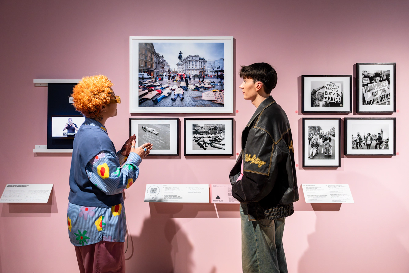 Photograph of 2 exhibition visitors in discussion in front of a pink coloured gallery wall on which 7 framed photographs are hung. The mix of black and white and colour photographs depict scenes of protest.