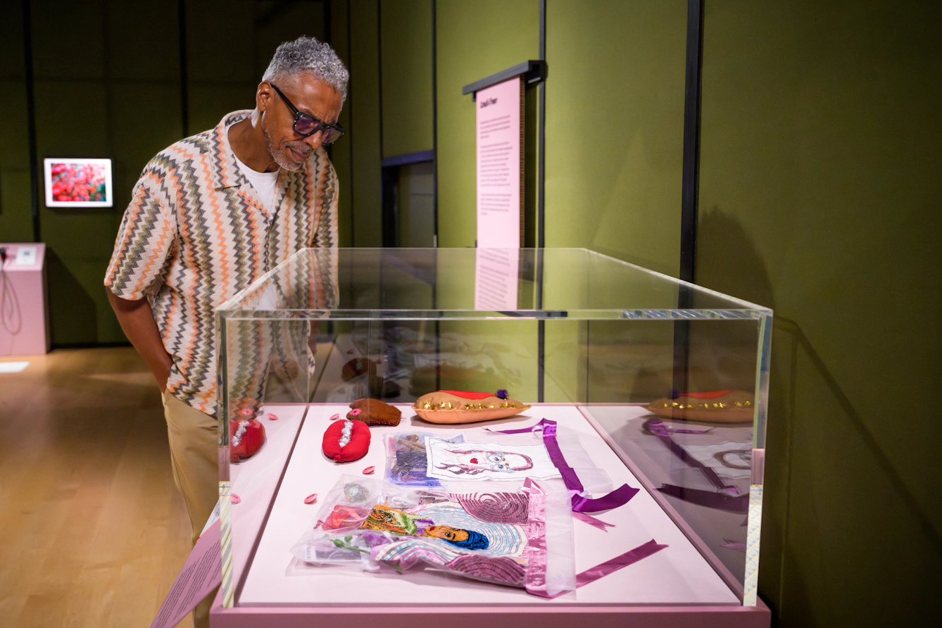 Photograph of an exhibition visitor looking into a display case containing multiple items, including several hand-made stitched items.