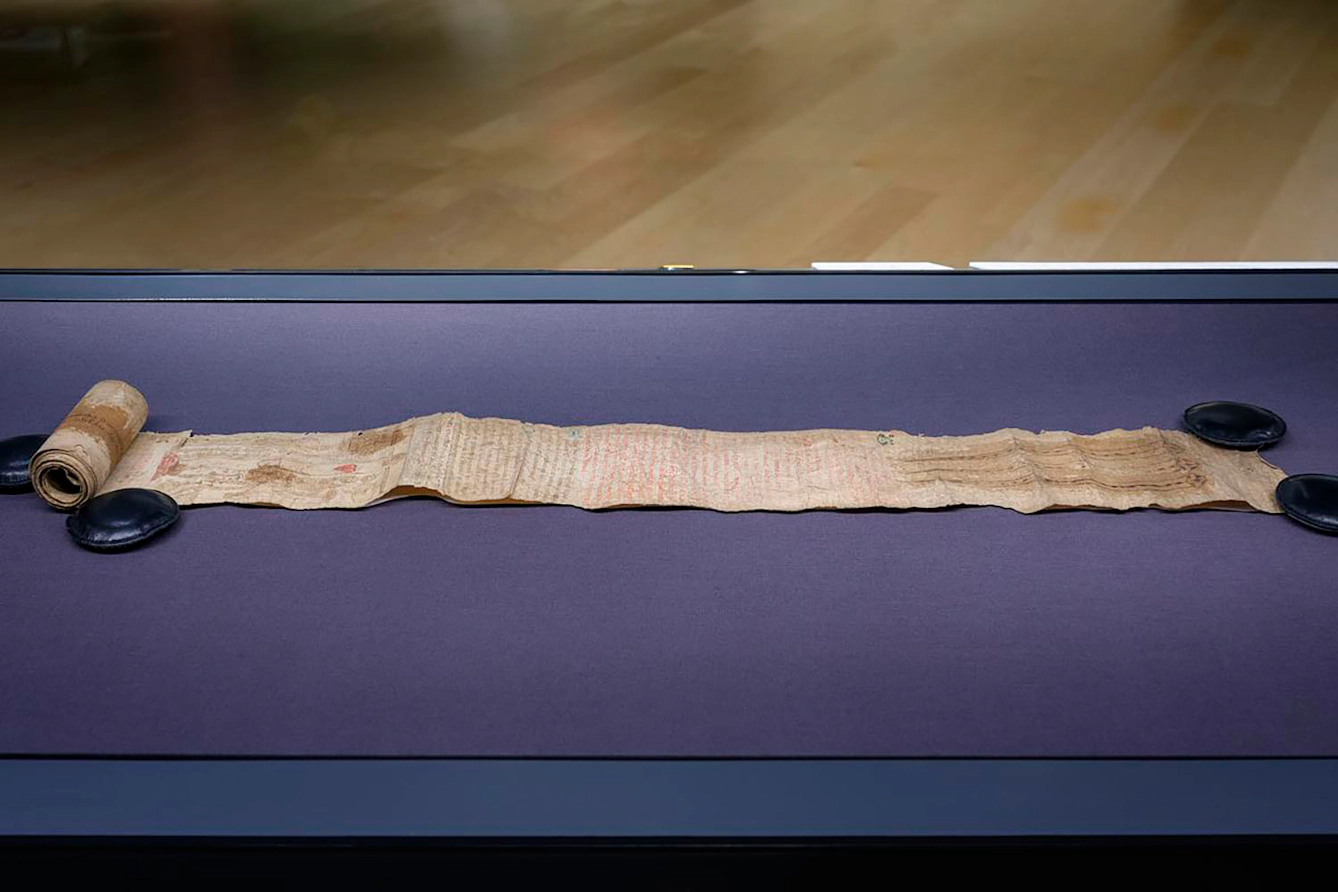 A partially unrolled medieval scroll made of parchment. The scroll is in a glass display case and held open by the flat discs of archival weights.