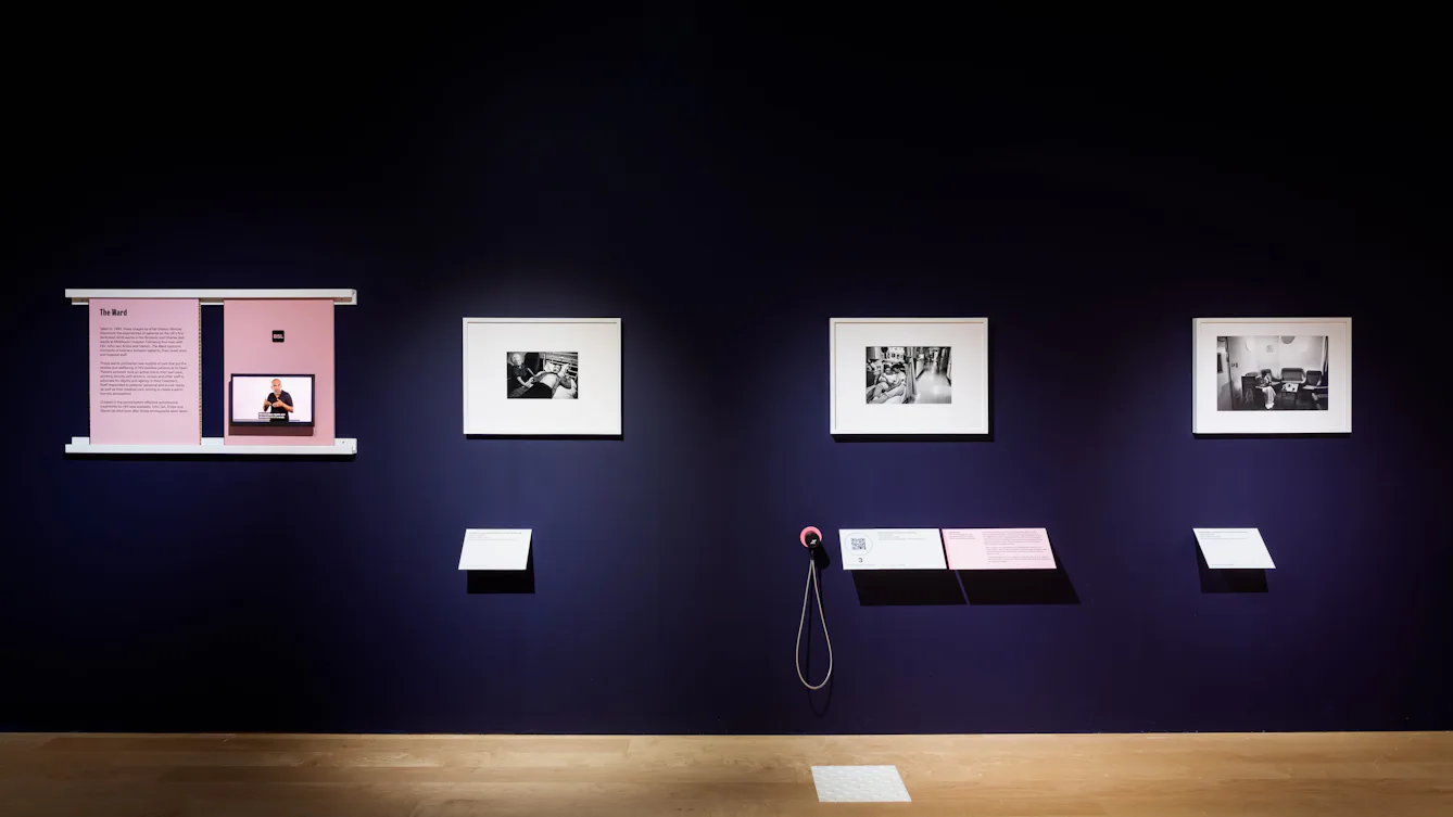 Wide view of a dark blue gallery wall displaying framed black-and-white photographs, with text panels and a small screen with a speaker, and audio handset below.