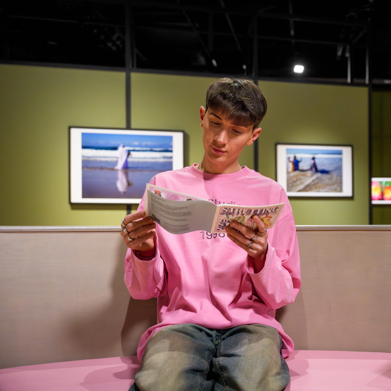 Visitor seated on a curved bench reads a leaflet in the gallery, with framed photographs on green walls in the background.