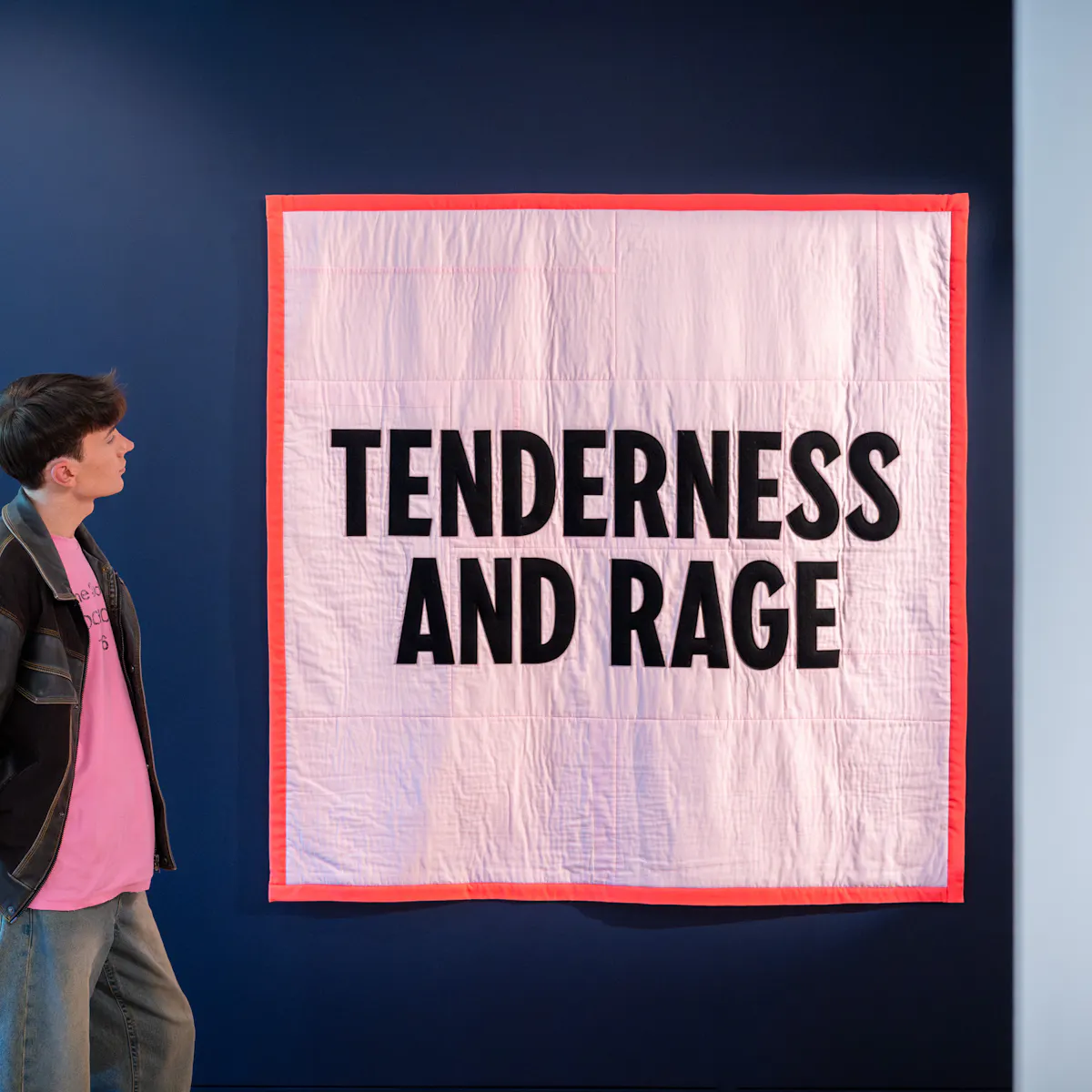 Photograph of a visitor standing at the entrance to an exhibition, looking at a large square quilt hanging on the wall. The quilt is light pink with a bright red hem, and has the words 'Tenderness and Rage' in capital letters sown onto it.