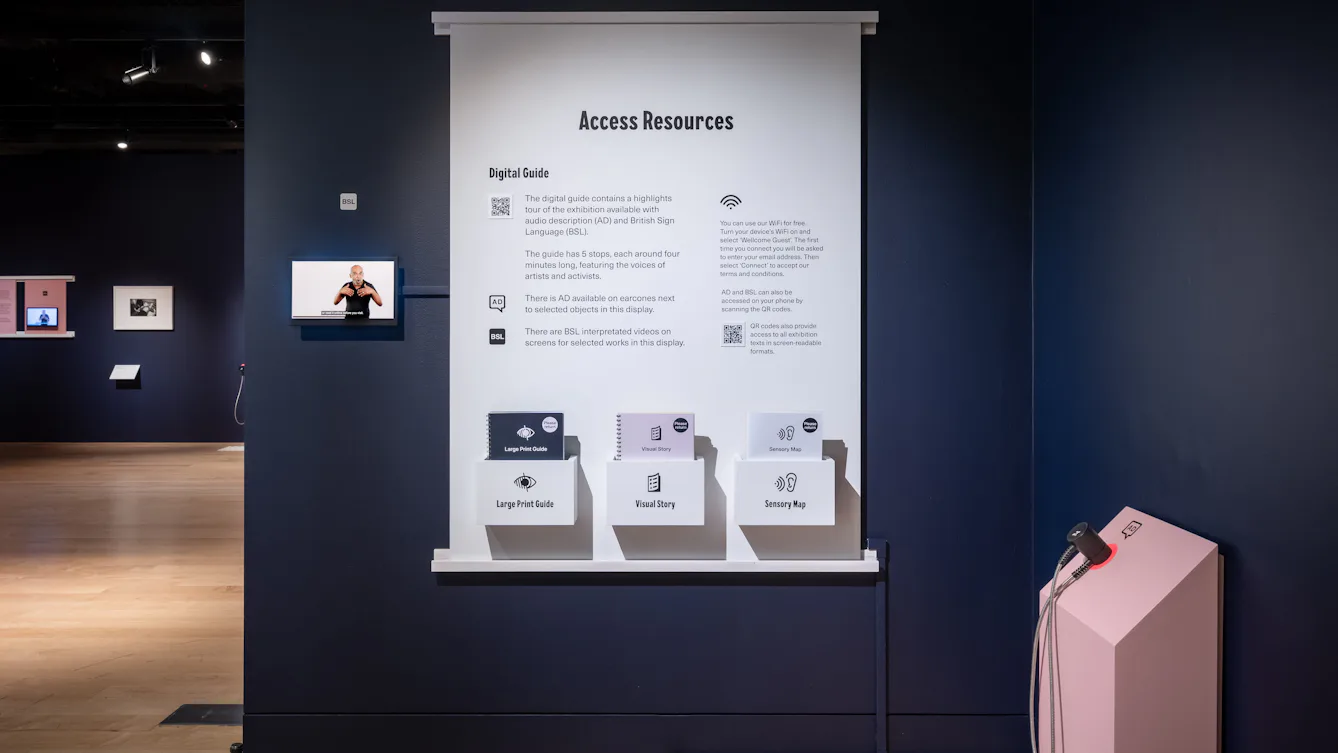 Display panel titled “Access Resources” with information about a digital guide, alongside shelves holding large print guides, visual stories and sensory maps, with a screen showing a BSL interpreter also on the wall.
