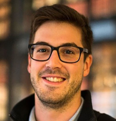 A black and white photo of the head and shoulders of a young man with dark hair and square-framed glasses. He is smiling and looking directly at the camera.
