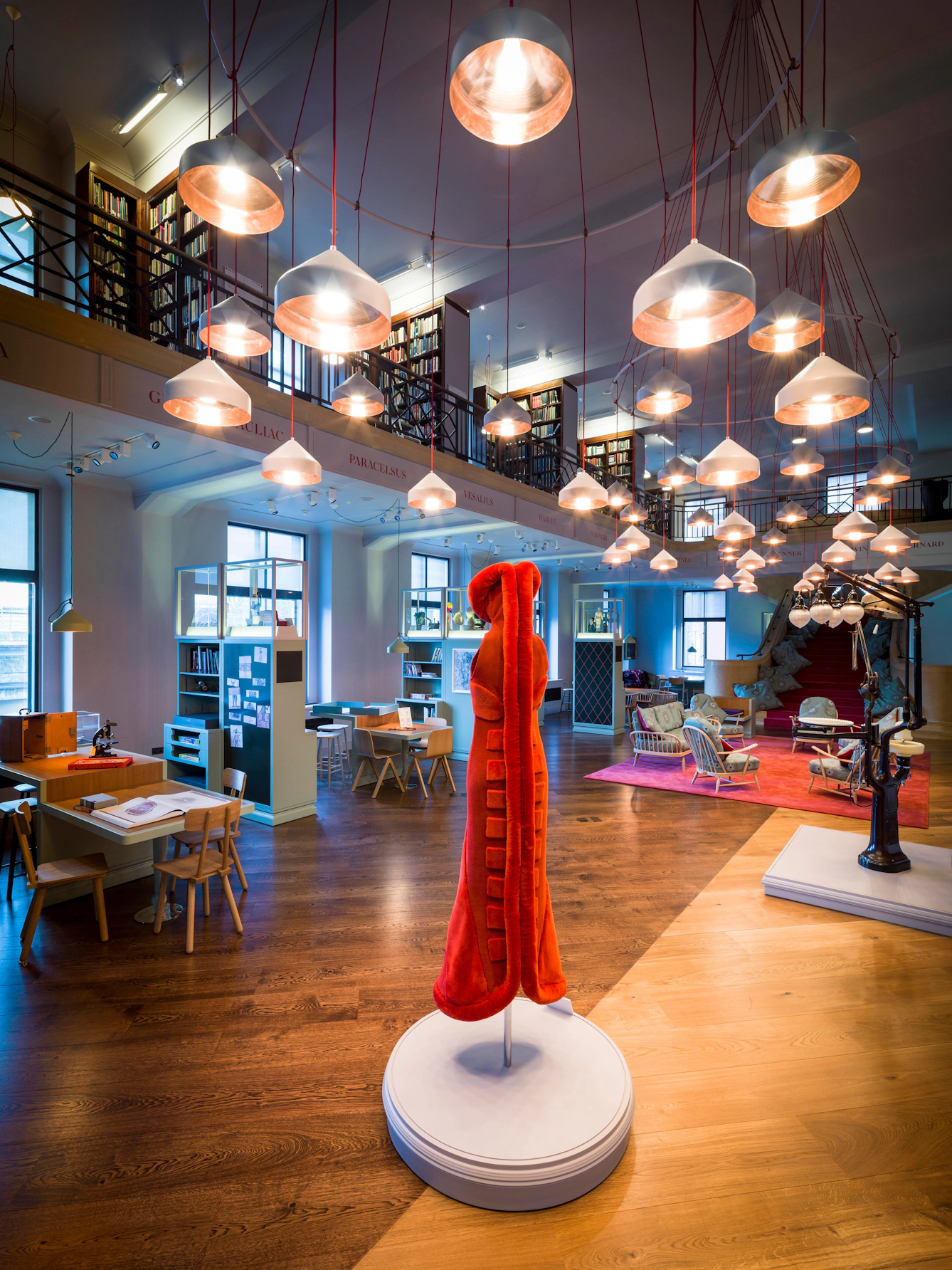 Open library and gallery space with display cases, reading tables and a central red sculptural figure, beneath rows of hanging pendant lights and a mezzanine lined with bookshelves.