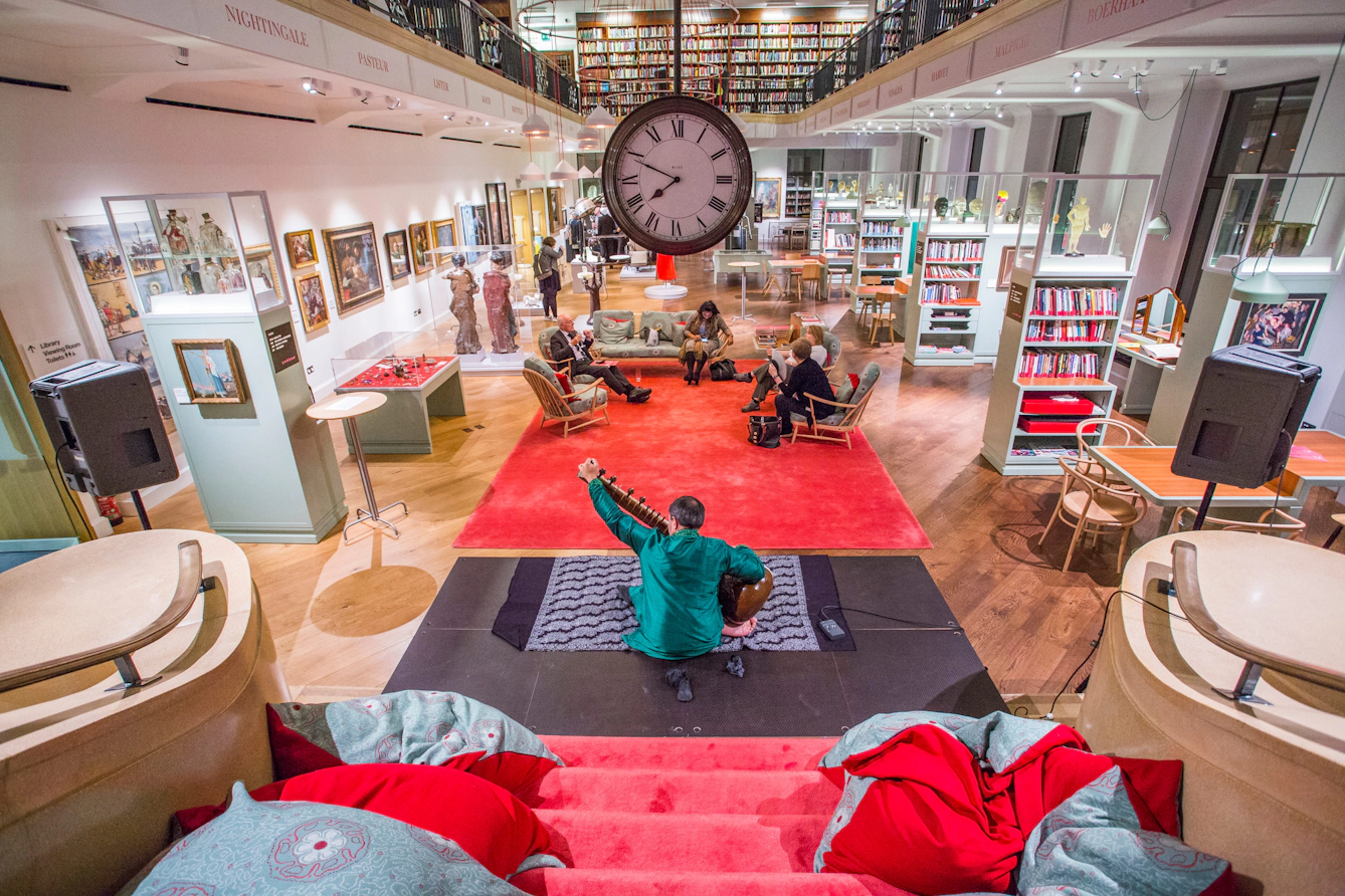 Library and gallery space with visitors seated around a red carpet and a musician performing beneath a large central clock and surrounding bookshelves.