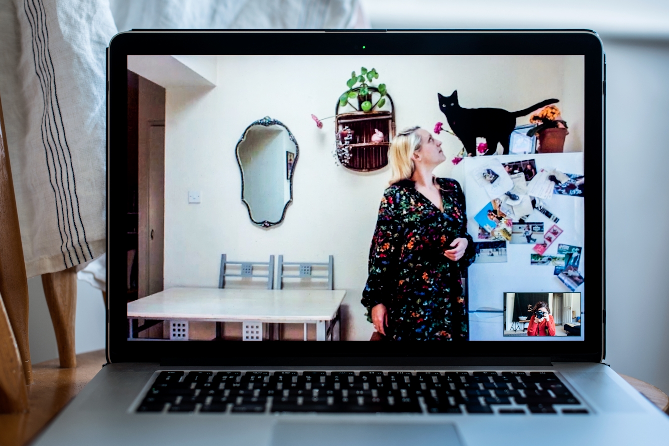 Photograph of an open laptop within a domestic scene. Most of the image is taken up with the screen, with part of the keyboard and trackpad visible. On the screen is a video call showing a woman standing in a kitchen setting, next to a fridge freezer. She is looking up to the top of the fridge where a black cat is standing looking towards the camera. In the bottom right corner of the screen the photographer can be seen in a small floating window, camera to her eye, in the process of taking the picture.