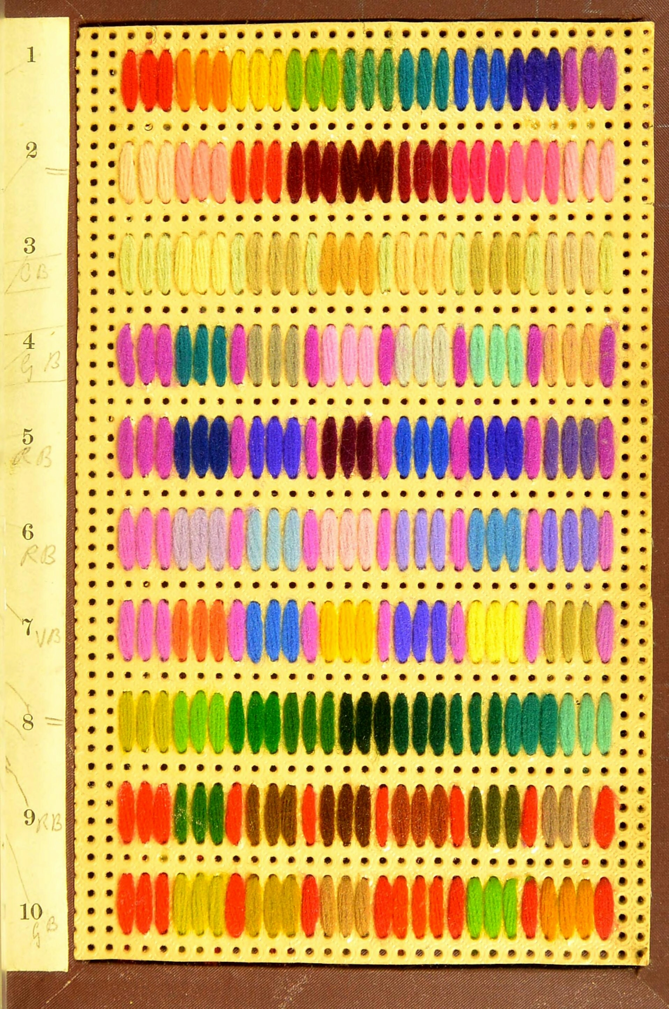A yellow card pierced with a grid of small holes. Different coloured wool yarns are threaded through the holes in 10 rows. The colours show gradual variations of e.g. green yarns in one row and conrasts of red, yellow and orrange yarns in another.