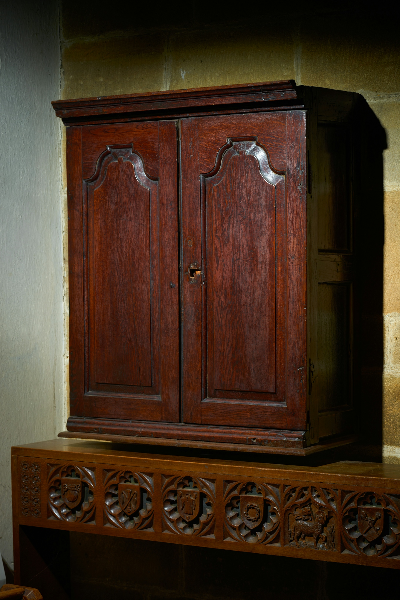 Photograph of a wooden cupboard with inlaid doors against the stone brick wall of a church.