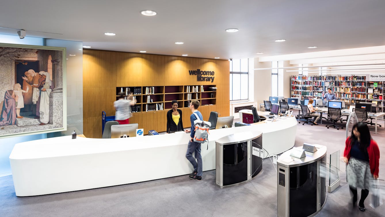 Photograph of the entrance to a library. In the centre is a large white information desk in the shape of a large curve. To the right are the security barriers which a lady in the process of exiting. A woman stands behind the information desk talking with a library visitor who is wearing a rucksack. Behind the desk someone is placing books on shelves and there is a large sign with the words, Wellcome Library. In the distance to the right the library shelves and visitors can be seen. To the far left is a section of a large oil painting, hung on a frosted glass wall.