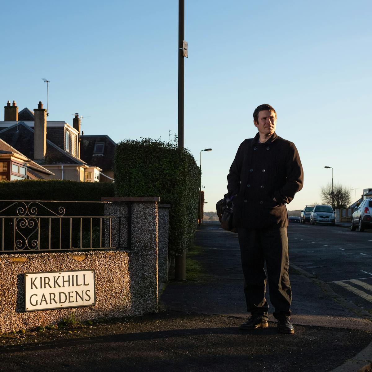 Photograph of a man in a black coat and trousers with a bag over his right shoulder. He is stood on a street corner, facing the camera but looking off to the right into the distance. A shaft of low golden sunlight picks him out from the suburban street scene around him made up of parked cars, houses and street lights. The sky is a pure blue. On a wall to the left is a road name sign carrying the words, 'Kirkhill Gardens'.