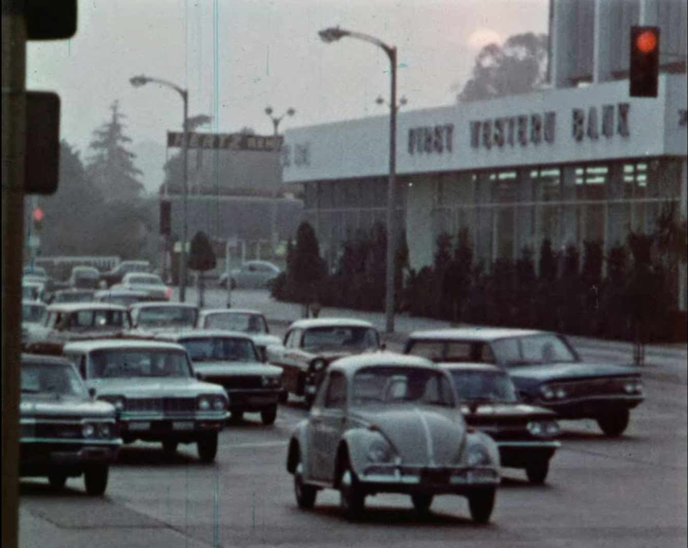 Film still showing a road filled with many late 1960s era cars driving along a road with a hazy sky in the background. 