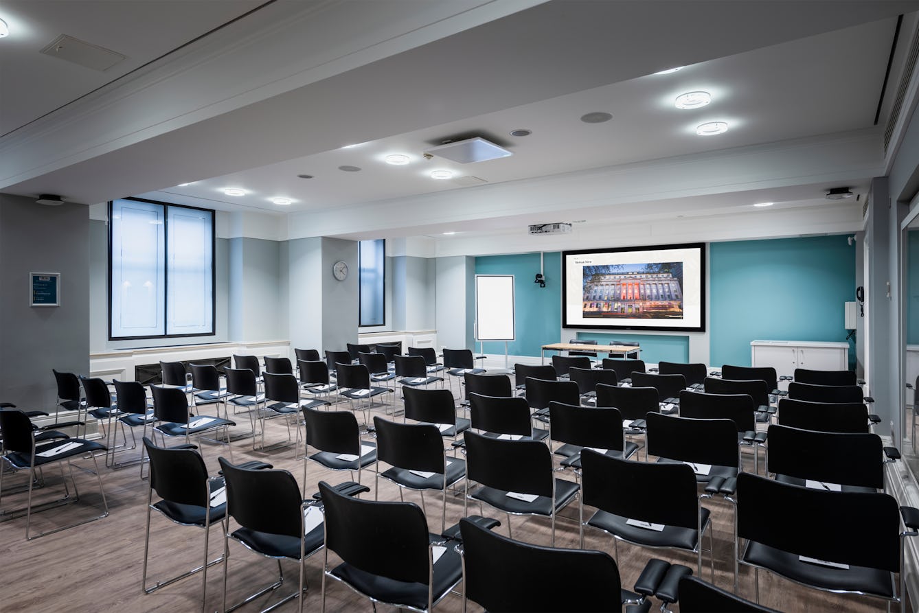 Photograph of the Franks and Steel room at the Wellcome Collection. 

Photograph shows a theatre space with chairs and a projector and screen set-up. There are large windows in the room. 
