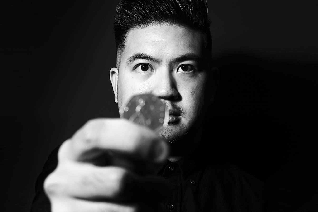 Black and white photographic portrait of a man dressed in a black shirt holding out a 50 pence piece to his the camera in his right hand. He is pictured from the chest up, his eyes directed towards the camera. The man's face and hand is spotlit in a small circle of light. He is standing against a black background which means he is surrounded by darkness.