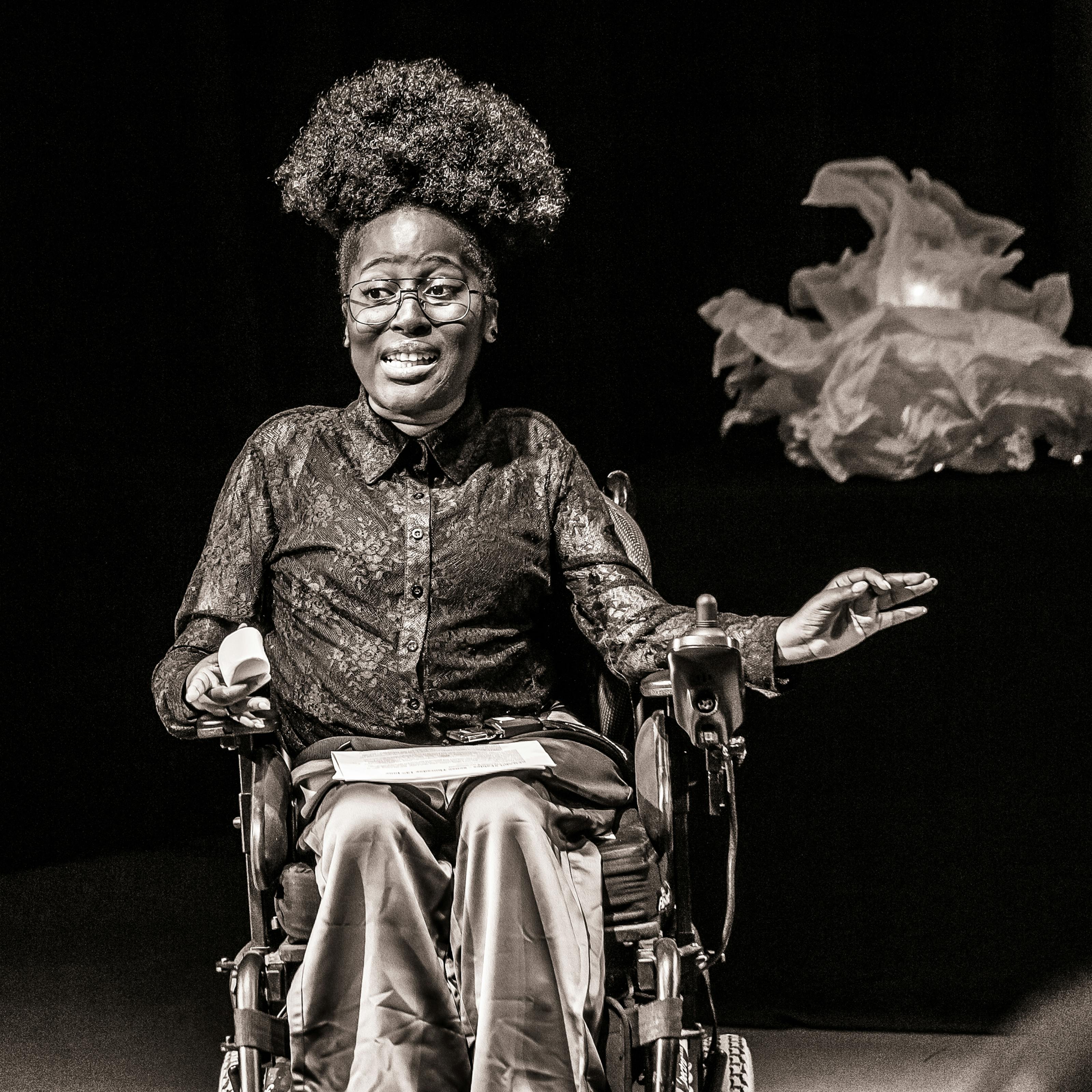 Black and white photograph with a warm tone. The image shows a young woman seated in a wheelchair against a black curtain and lit by a spotlight. She is performing to an audience. She has been captured 'in action'.