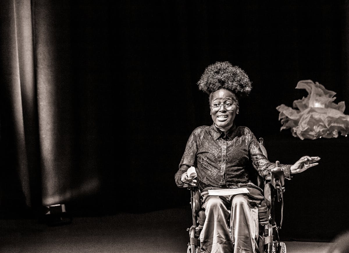 Black and white photograph with a warm tone. The image shows a young woman seated in a wheelchair against a black curtain and lit by a spotlight. She is performing to an audience. She has been captured 'in action'.