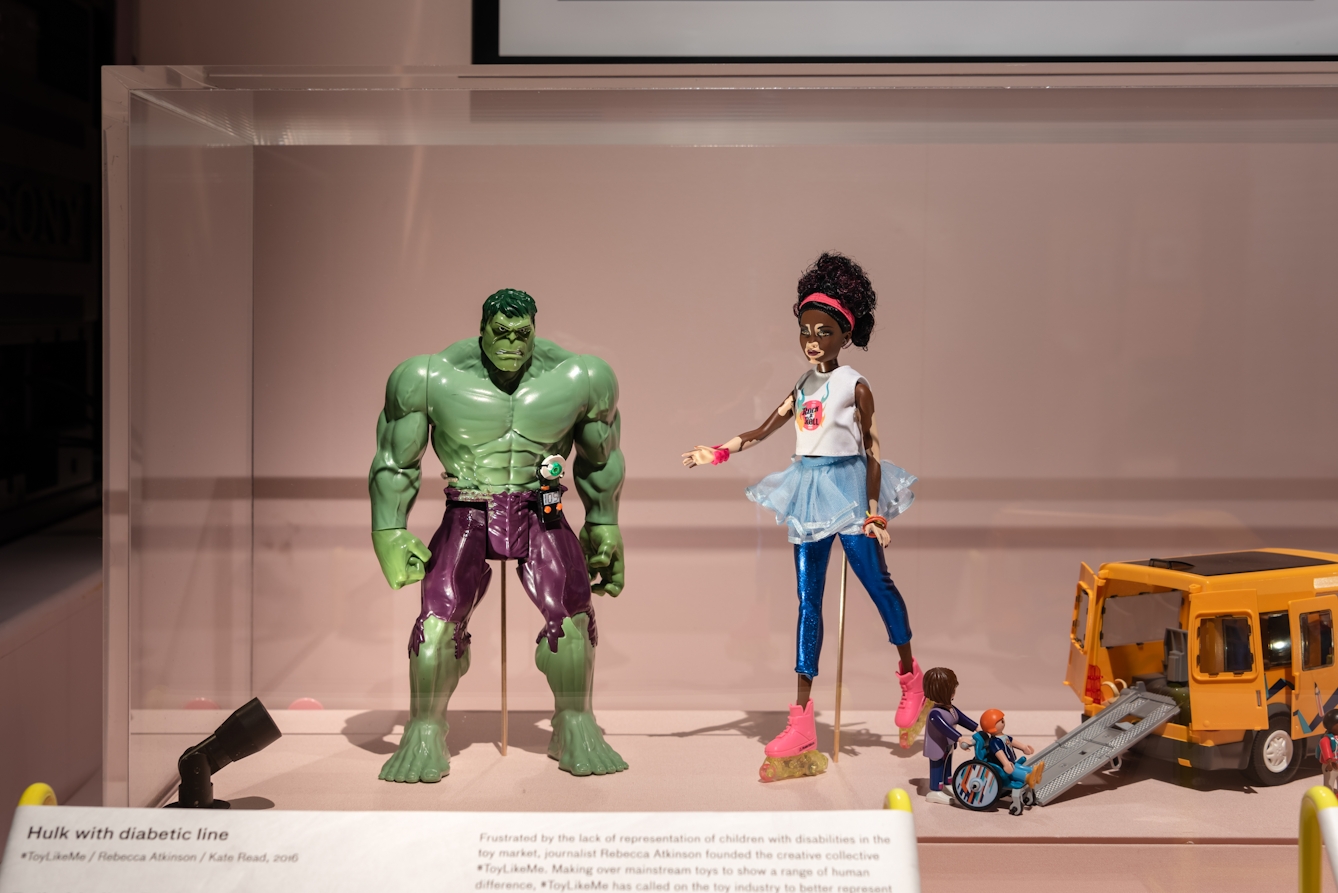 Photograph of an exhibition display case showing from left to right, an Incredible Hulk toy with a diabetic medication line attached, a Barbie doll with vitiligo and a Playmobile vehicle with extended rear access ramp and a child in a wheelchair.