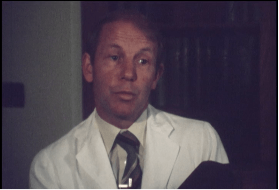 Film still of a man wearing a tie and white doctor's coat.
