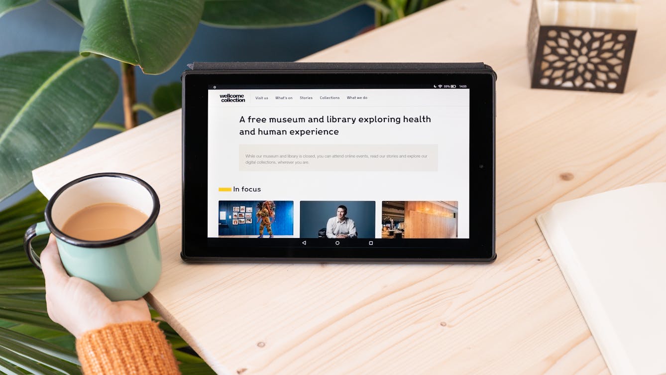 Photograph of a tablet standing on a table showing Wellcome Collection's website on the screen. The web page shows the text "A free museum and library exploring health and human experience". A hand holding a mug of tea is next to the tablet.