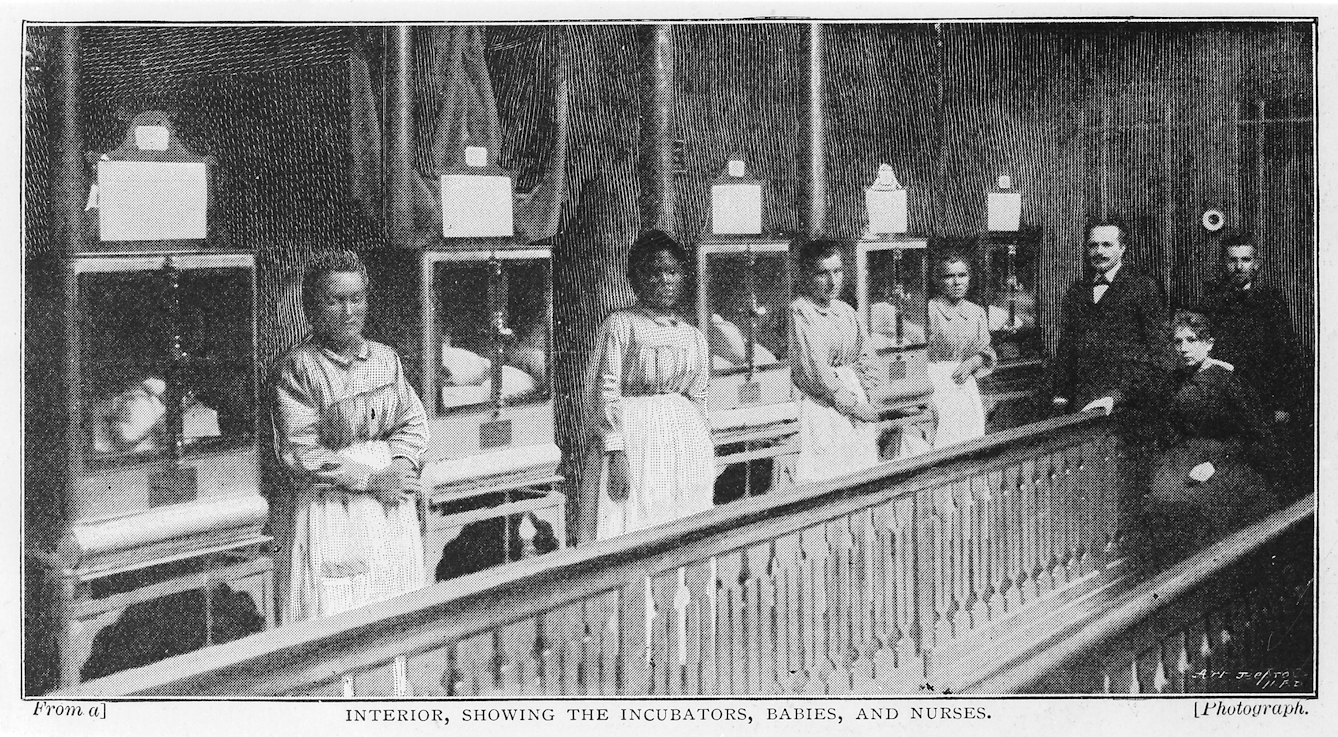 Black and white photograph from 1896 showing a row of five early incubators, each containing a baby, attended by four female nurses in uniform. There are three other people in the image, wearing formal clothing - two men and a woman.