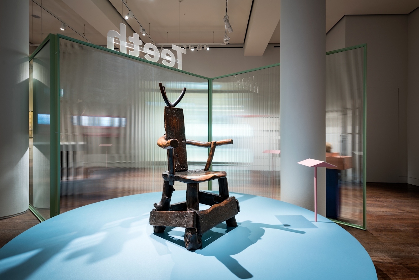 Photograph of a wooden barber-surgeon's chair on a blue plinth in the Teeth exhibition at Wellcome Collection.