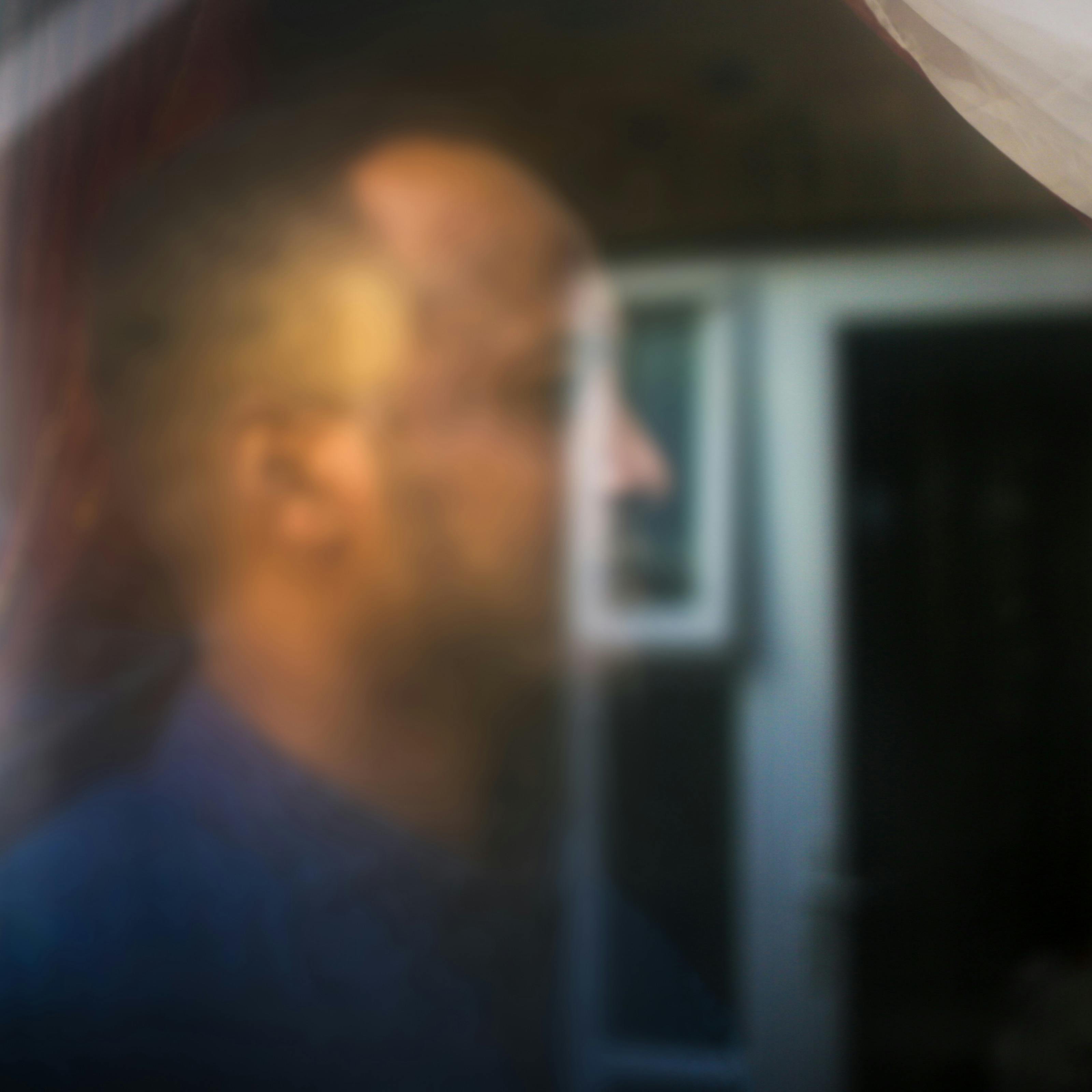 Photograph of a man's head seen through a glass window. His head is out of focus and merges with the reflection of another scene.