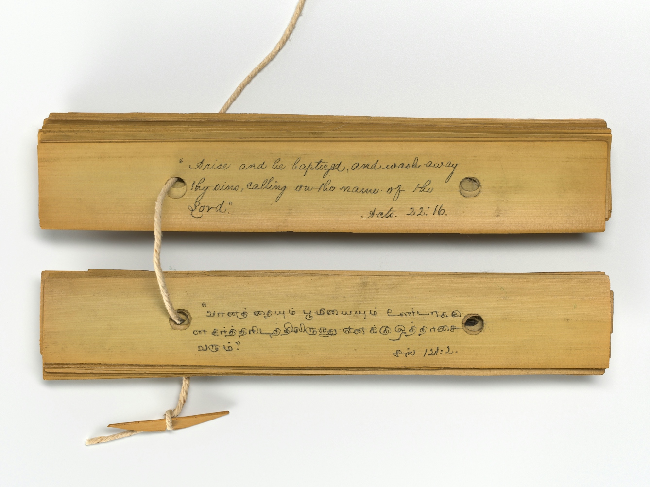 An open palm leaf manuscrip showing two pages, one in English and one in Tamil. The leaves of the manuscript and connected by a string passing through a hole piercing all of the leaves. The end of the string is fastened to a small wooden peg. The English text says 'Arise and be baptized and wash away the sins calling out the names of the Lord. Acts 2.2:16'
